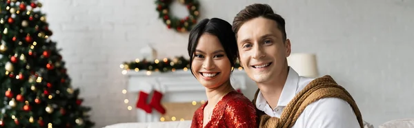 Jovem casal multiétnico sorrindo para a câmera perto de decoração de Natal borrada em casa, banner — Fotografia de Stock