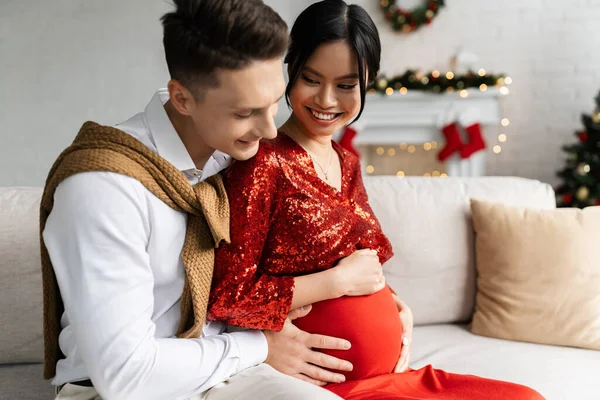 Schwangere und elegante Asiatin lächelt in der Nähe Ehemann berührt ihren Bauch auf Sofa im Wohnzimmer mit Weihnachtsdekor — Stockfoto