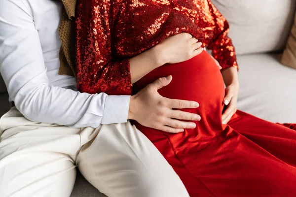 Vista parcial de la mujer embarazada en ropa elegante roja sentada cerca del marido abrazando su vientre - foto de stock