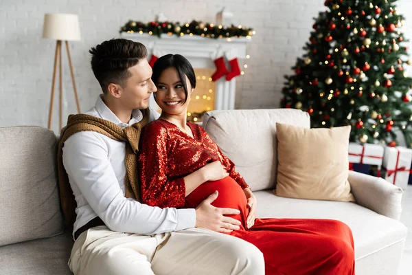 Felice uomo che abbraccia pancia di incinta moglie asiatica in abiti festivi sul divano in soggiorno con arredamento natalizio — Foto stock