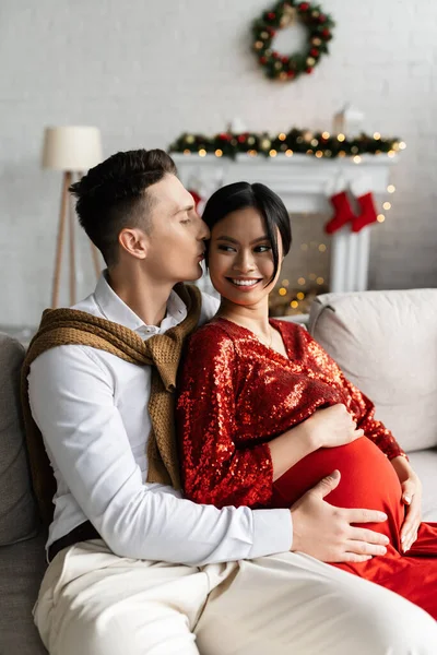 Hombre besando embarazada asiática mujer y abrazando su vientre durante la celebración de la Navidad - foto de stock