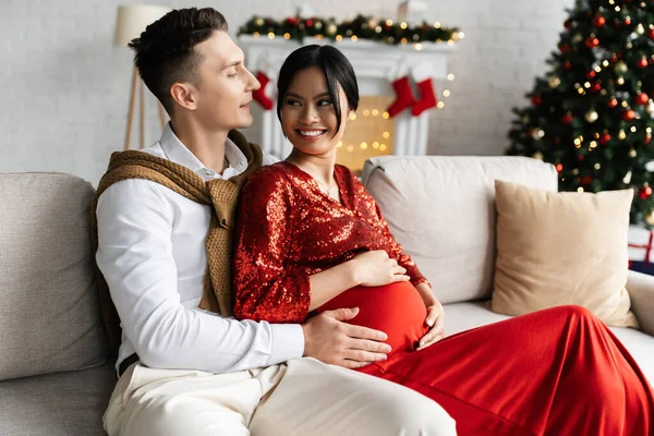 Giovane uomo che abbraccia pancia di incinta ed elegante moglie asiatica sul divano in soggiorno con arredamento natalizio — Foto stock