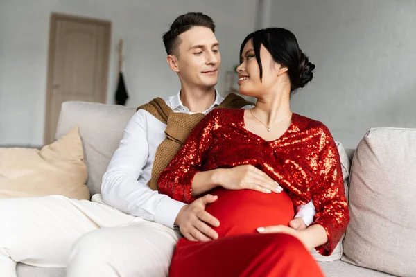 Incinta asiatico donna in rosso e lucido vestiti sorridente a giovane marito abbraccio il suo pancia su divano a casa — Foto stock