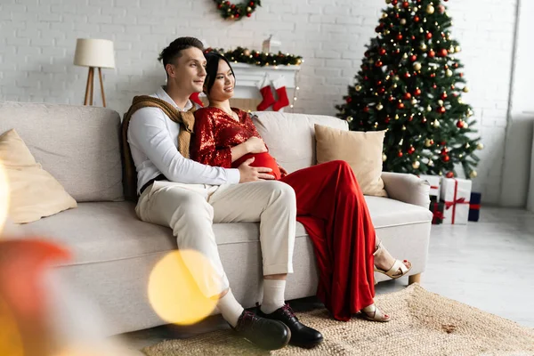 Pregnant asian woman in elegant clothes sitting with husband on sofa in living room near Christmas tree — Stock Photo