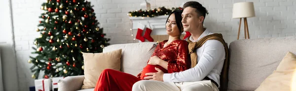 Hombre joven y mujer asiática embarazada en ropa festiva en acogedor sofá en la sala de estar con árbol de Navidad, pancarta - foto de stock