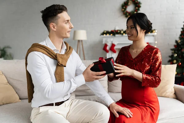 Junger Mann präsentiert Weihnachtsgeschenk an schwangere und elegante asiatische Frau, während er zu Hause auf der Couch sitzt — Stockfoto