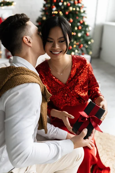 Joven hombre celebración Navidad regalo y besos embarazada asiático mujer en elegante ropa - foto de stock