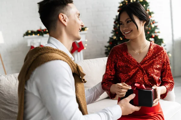 Schwangere Asiatin lächelt Ehemann an, der Weihnachtsgeschenk auf Sofa zu Hause präsentiert — Stockfoto