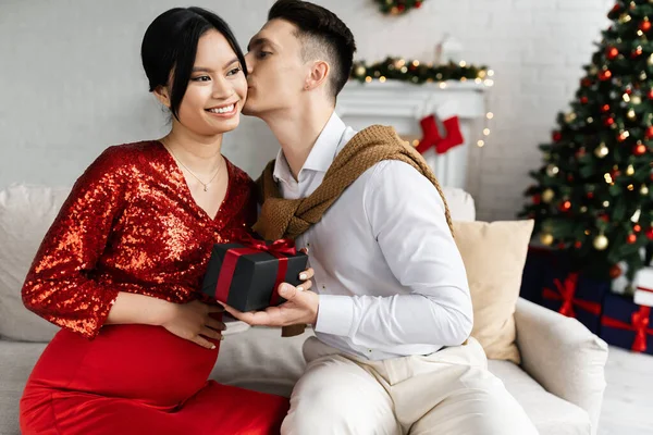 Grávida asiático mulher no elegante roupas sorrindo perto marido com Natal presente beijá-la no sofá em casa — Fotografia de Stock