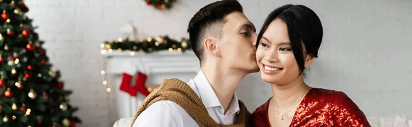 Brunette asian woman smiling near young husband kissing her cheek during Christmas celebration, banner — Stock Photo
