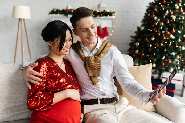 Homem feliz com ultra-som digitalização abraçando mulher asiática grávida em roupas elegantes perto borrada árvore de Natal — Fotografia de Stock