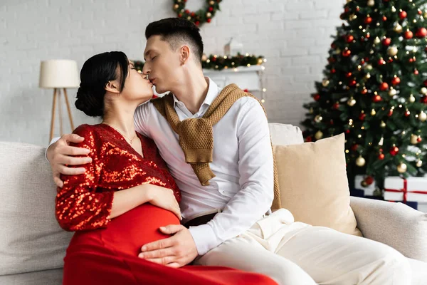 Мужчина целует беременную азиатку и обнимает ее животик на диване возле размытой рождественской елки — стоковое фото