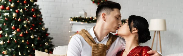 Young man kissing brunette asian wife near Christmas tree on blurred background, banner — Stock Photo