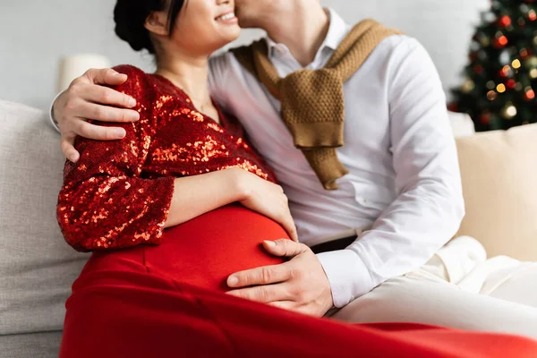 Cropped view of man embracing pregnant wife in red and shiny clothes while celebrating Christmas at home — Stock Photo