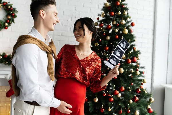 Felice ed elegante asiatico donna holding ultrasuoni scansione vicino felice marito e albero di Natale — Foto stock