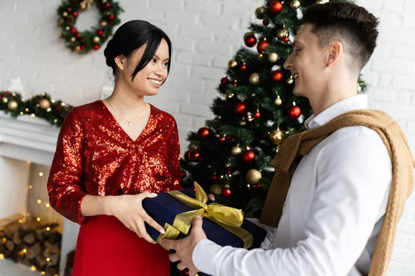 Embarazada y complacido asiático mujer tomando regalo caja de marido cerca decorado árbol de Navidad - foto de stock