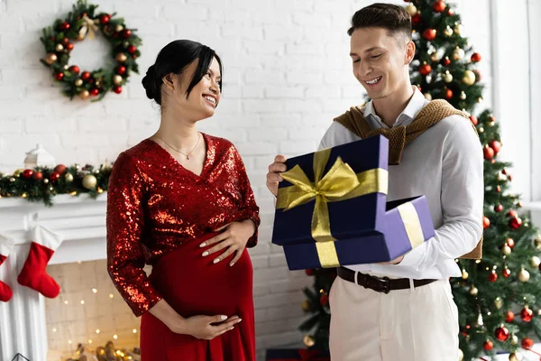 Incinta asiatica donna sorridente vicino felice marito apertura regalo scatola in soggiorno con decorazioni natalizie — Foto stock
