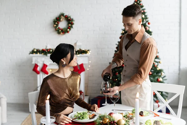 Homem elegante derramando vinho perto asiático esposa durante romântico ceia no Natal celebração — Fotografia de Stock