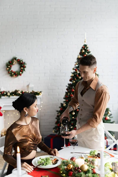 Elegant man pouring red wine near asian wife in festive clothes during romantic Christmas supper at home — Stock Photo