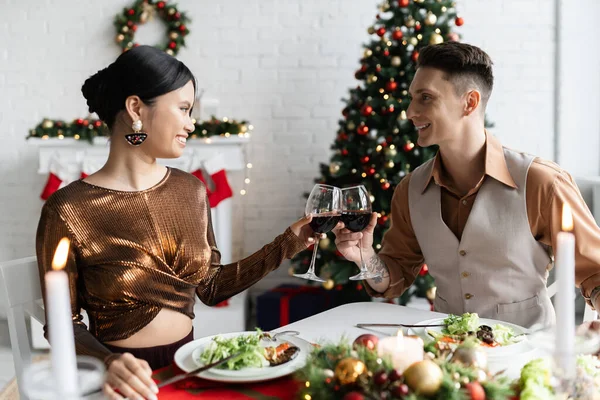 Весела міжрасова пара кмітливих келихів вина під час романтичної вечері у вітальні з різдвяним декором — стокове фото