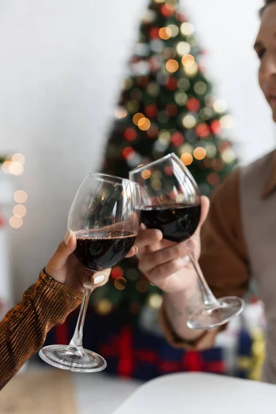 Vista recortada de la pareja joven tintineo copas de vino mientras se celebra la Navidad sobre fondo borroso - foto de stock