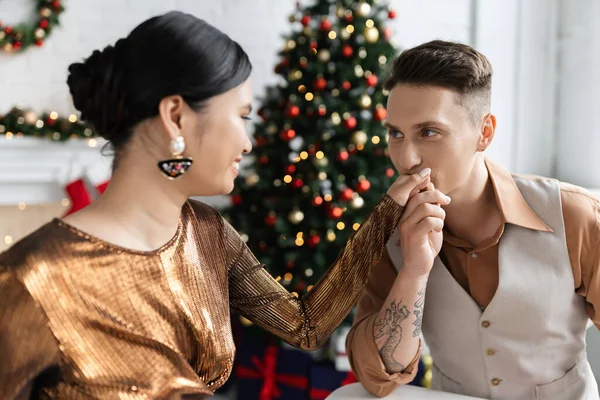 Tattooed man kissing hand of smiling asian wife in elegant shiny blouse — Stock Photo