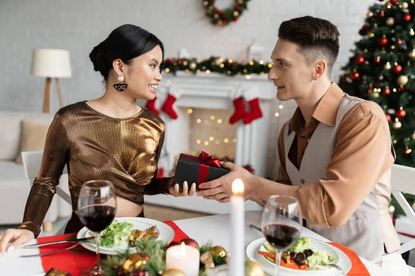 Junger Mann gibt Weihnachtsgeschenk an glückliche asiatische Frau beim romantischen Abendessen — Stockfoto