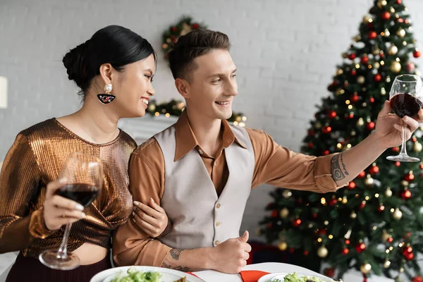 Homem sorrindo brindar com copo de vinho enquanto celebra o Natal com feliz esposa asiática — Fotografia de Stock