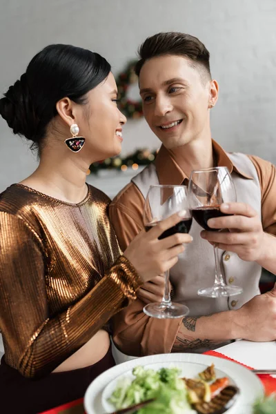 Радісна багатоетнічна пара в елегантному вбранні дивиться один на одного і клінкінг винних окулярів під час романтичної вечері — стокове фото