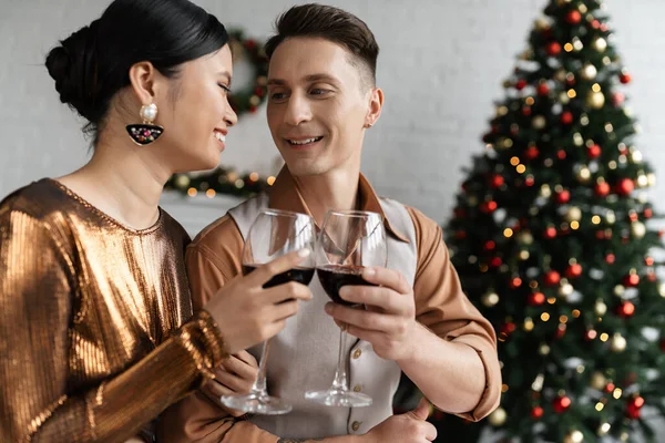 Glückliches multiethnisches Paar im eleganten Outfit, das neben verschwommenem Weihnachtsbaum Weingläser klimpert — Stockfoto