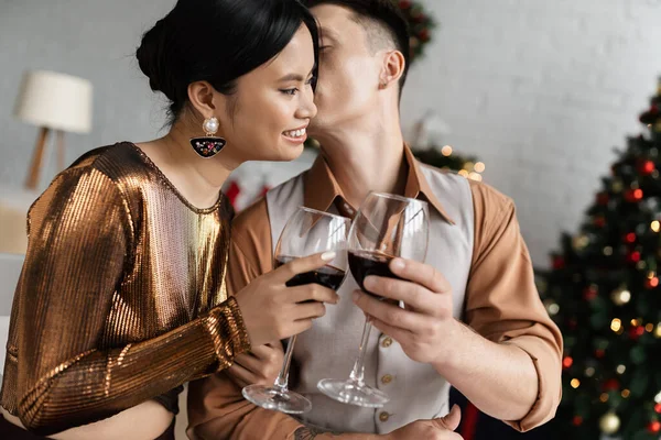 Young man kissing happy and elegant asian wife and clinking wine glasses while celebrating Christmas — Stock Photo