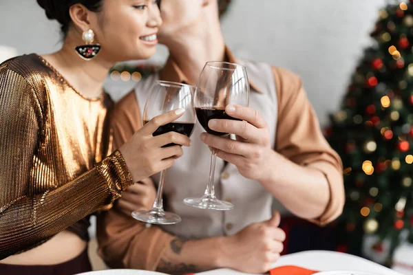 Sonriente asiático mujer en brillante blusa tintineando copas de vino con borrosa marido durante la celebración de Navidad — Stock Photo