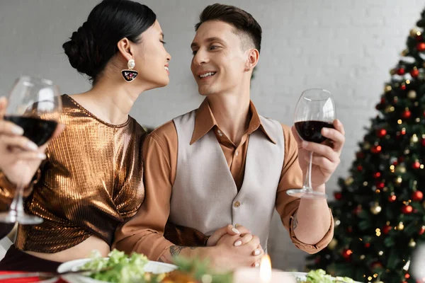 Межрасовая пара в праздничной одежде, держась за руки и улыбаясь друг другу, держа бокалы для вина — стоковое фото