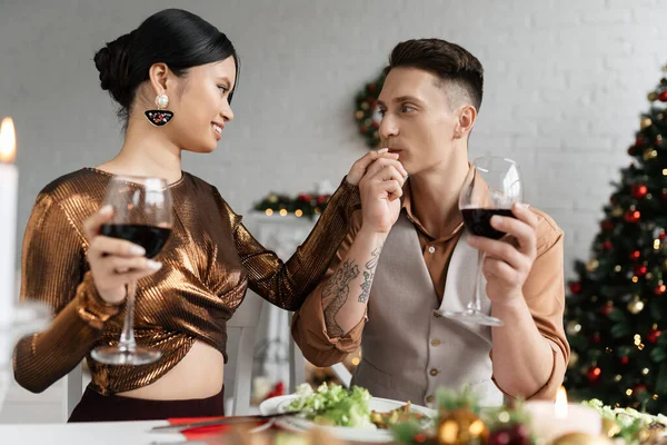 Tattooed man kissing hand of elegant asian wife while celebrating Christmas at home — Stock Photo