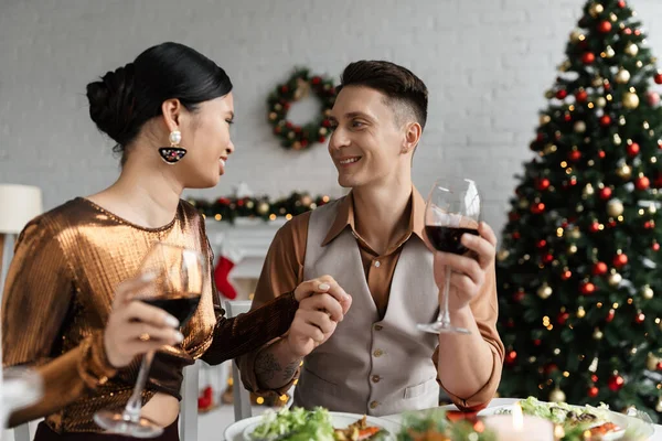 Elegante pareja interracial con copas de vino cogidas de la mano y sonriéndose durante la cena romántica de Navidad - foto de stock