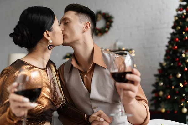 Jovem e elegante casal multiétnico com copos de vinho de mãos dadas e beijando durante a celebração de Natal — Fotografia de Stock
