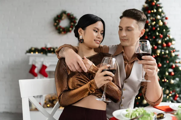 Interracial romantic couple holding hands and wine glasses during Christmas supper at home — Stock Photo