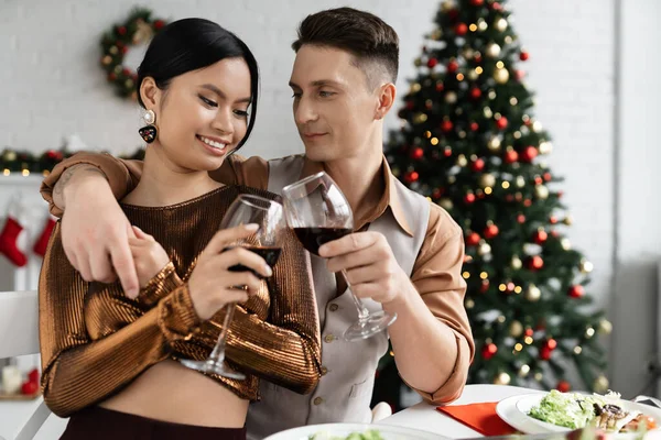 Улыбающийся мужчина обнимает элегантную азиатскую жену и звенит бокалами вина во время праздничного романтического ужина — стоковое фото