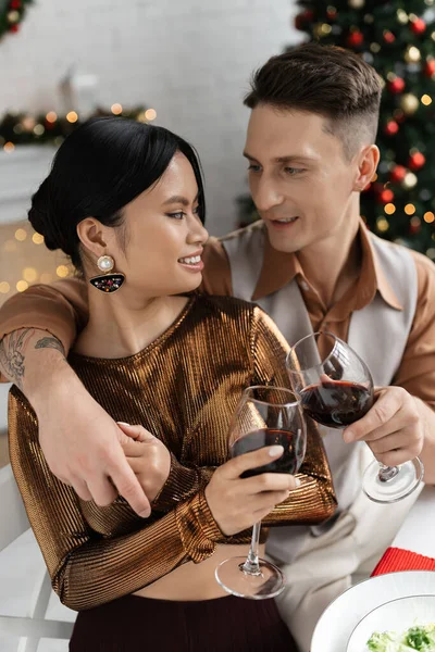 Elegante pareja multiétnica tintineo copas de vino durante la cena romántica festiva en casa - foto de stock