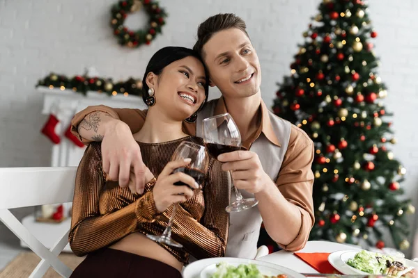 Cheerful and elegant multiethnic couple clinking wine glasses during Christmas supper at home — Stock Photo
