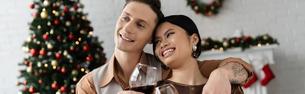 Heureux couple interracial cliquetis verres à vin près de l'arbre de Noël sur fond flou, bannière — Photo de stock