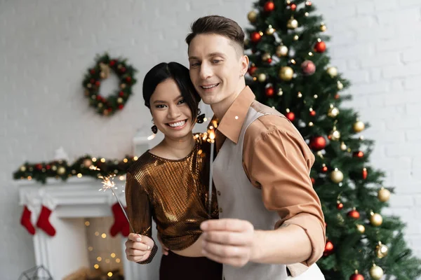 Animado casal multiétnico em roupa festiva segurando sparklers perto da árvore de Natal decorada no fundo borrado — Fotografia de Stock