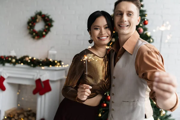 Cheerful and elegant interracial couple holding shiny sparklers and looking at camera during Christmas celebration — Stock Photo