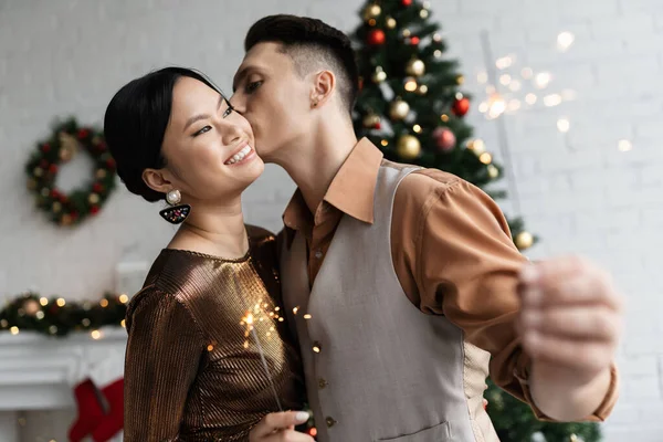 Young man kissing cheek of excited asian wife near shiny sparklers during Christmas celebration — Stock Photo
