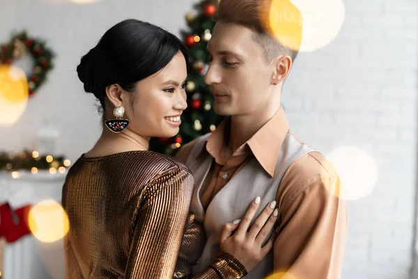 Morena asiática mulher em roupas festivas sorrindo perto de jovem marido durante a celebração de Natal em casa — Fotografia de Stock