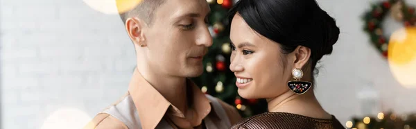 Felice uomo guardando allegro ed elegante moglie asiatica durante la celebrazione di Natale, banner — Foto stock