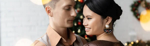 Happy man looking at cheerful and elegant asian wife during Christmas celebration, banner — Stock Photo