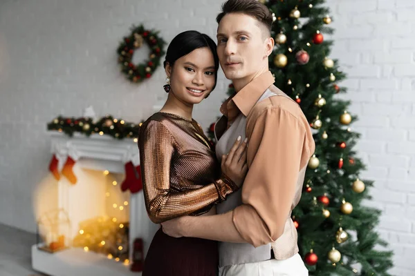 Jovem e elegante casal interracial abraçando e sorrindo para a câmera perto de árvore de Natal borrada — Fotografia de Stock