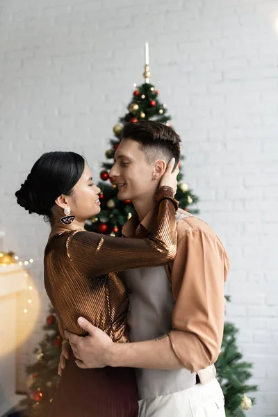 Homem feliz e sorrindo mulher asiática em roupas festivas abraçando perto da árvore de Natal — Fotografia de Stock