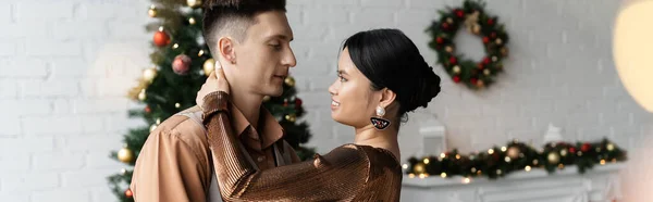 Giovane coppia interrazziale in abiti festivi abbracciandosi vicino all'albero di Natale, striscione — Foto stock